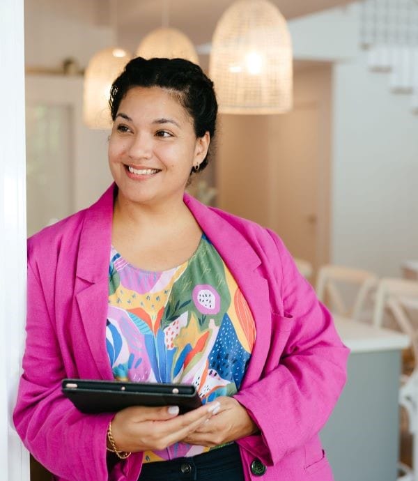 Brown woman with black hair looks to the left, smiling. She wears pink blazer with multicoloured handmade top. Kira Hansen provides social media and website services for women in business at Brisbane Creative Web Studio.
